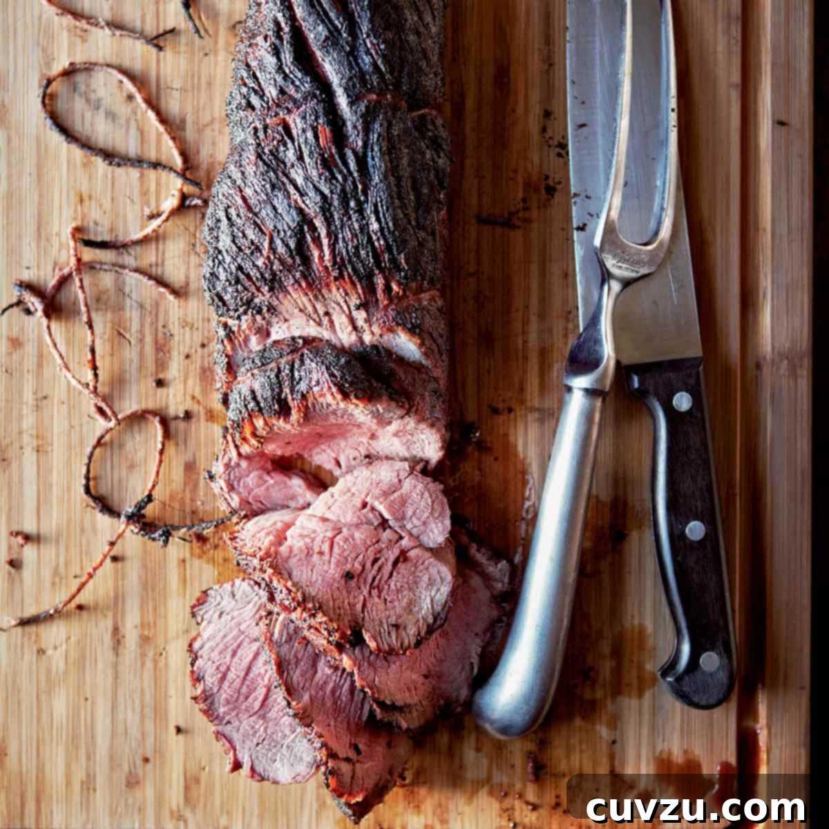 A whole grilled beef tenderloin, partially sliced on a wooden cutting board with a knife, meat fork, and twine beside it.