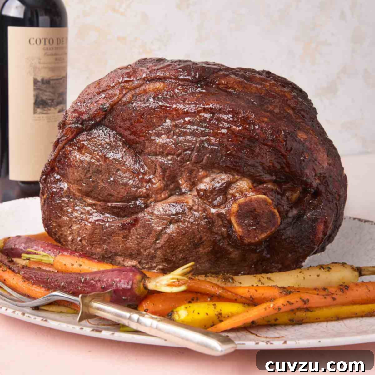 A roasted fresh ham on a platter with roasted carrots and a bottle of red wine in the background.