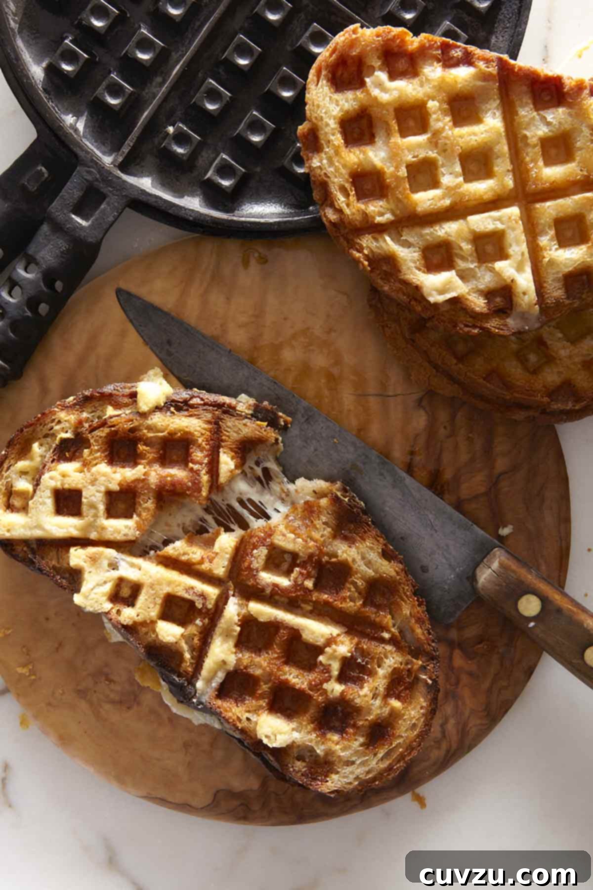 Two waffle iron grilled cheese sandwiches on a wooden cutting board with a knife and a waffle iron nearby.