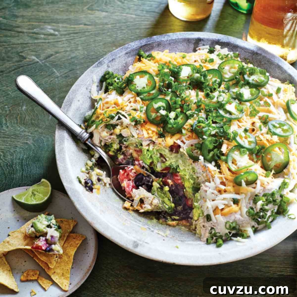 A bowl filled with seven-layer dip with a spoon resting inside and a plate with tortilla chips, dip, and a lime wedge on the side.