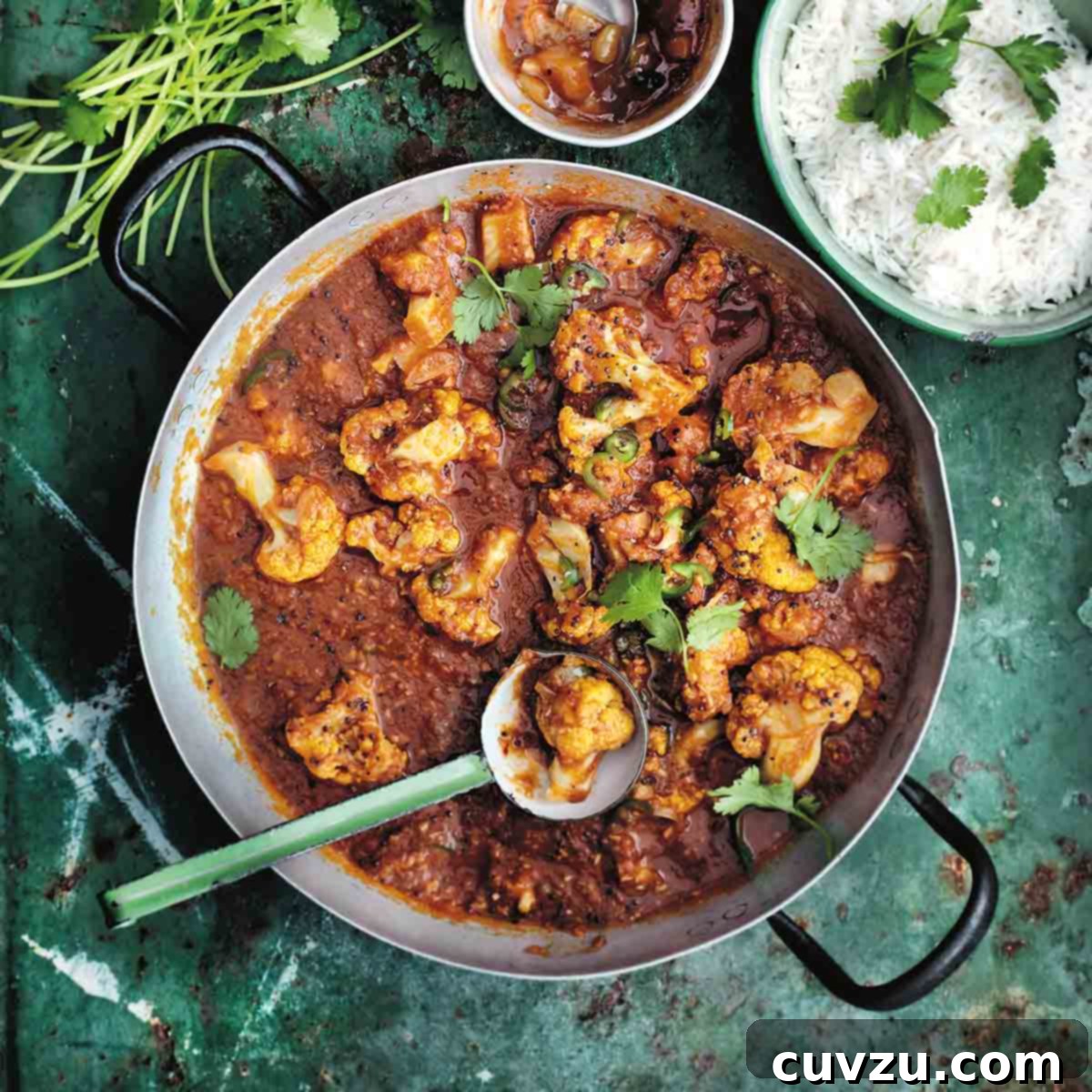 A metal pot filled with fragrant cauliflower and tomato curry with a ladle resting inside, accompanied by a bowl of rice and chutney.