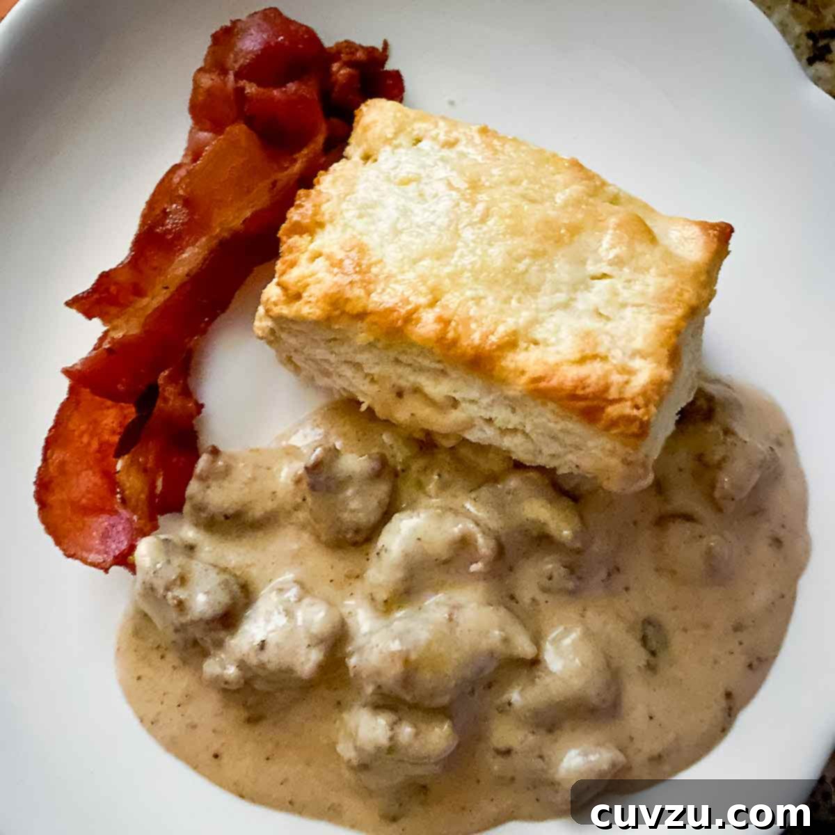 A white plate showcasing a large Cat Head biscuit, split and generously topped with creamy sausage gravy, alongside two crispy strips of bacon.