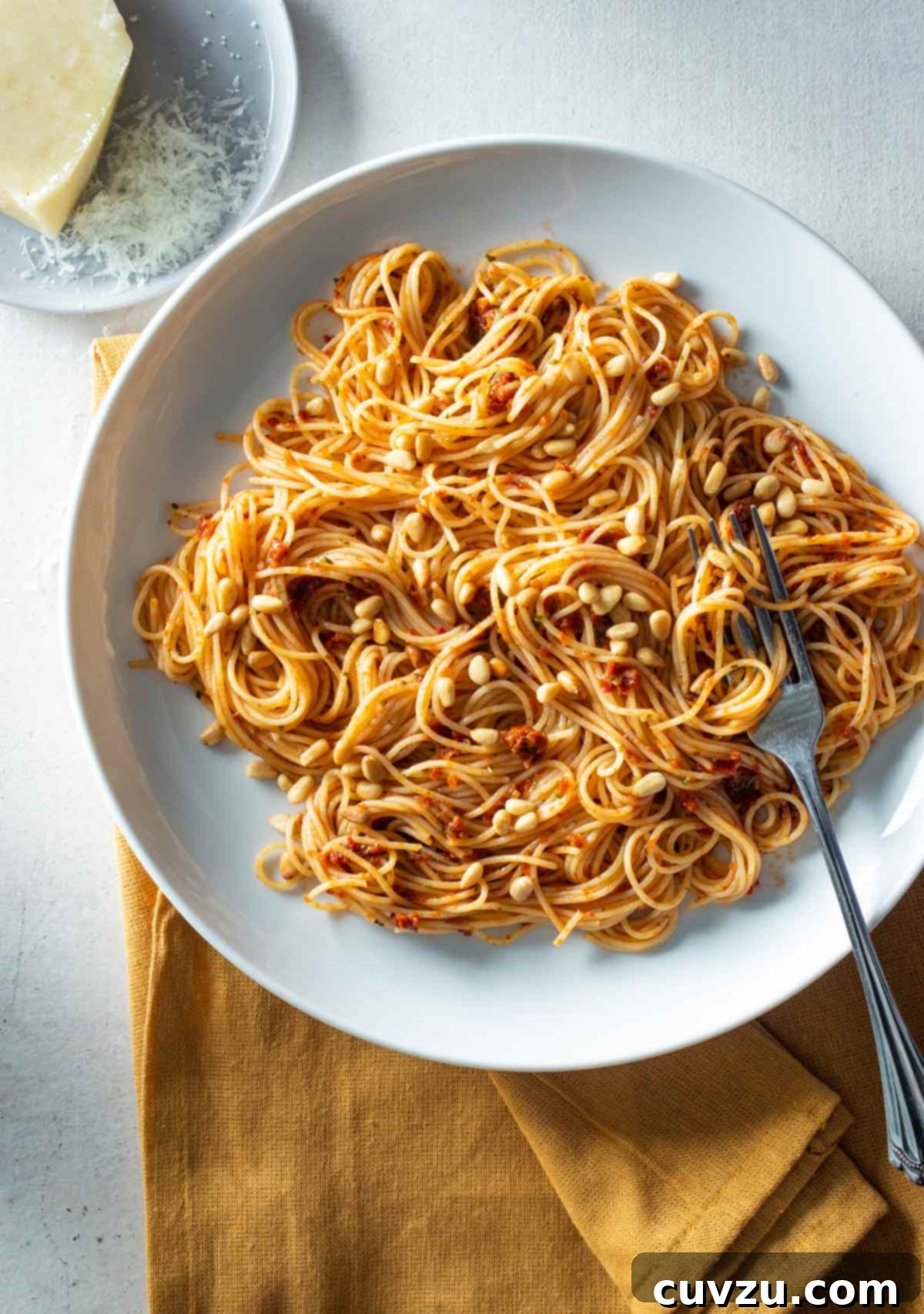 Vibrant Sun-Dried Tomato Pasta 2 A white bowl filled with sun-dried tomato pasta and pine nuts with a fork resting inside and a block of parmesan on a plate beside it.