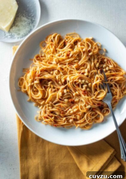 A white bowl filled with sun-dried tomato pasta and pine nuts with a fork resting inside and a block of parmesan on a plate beside it.