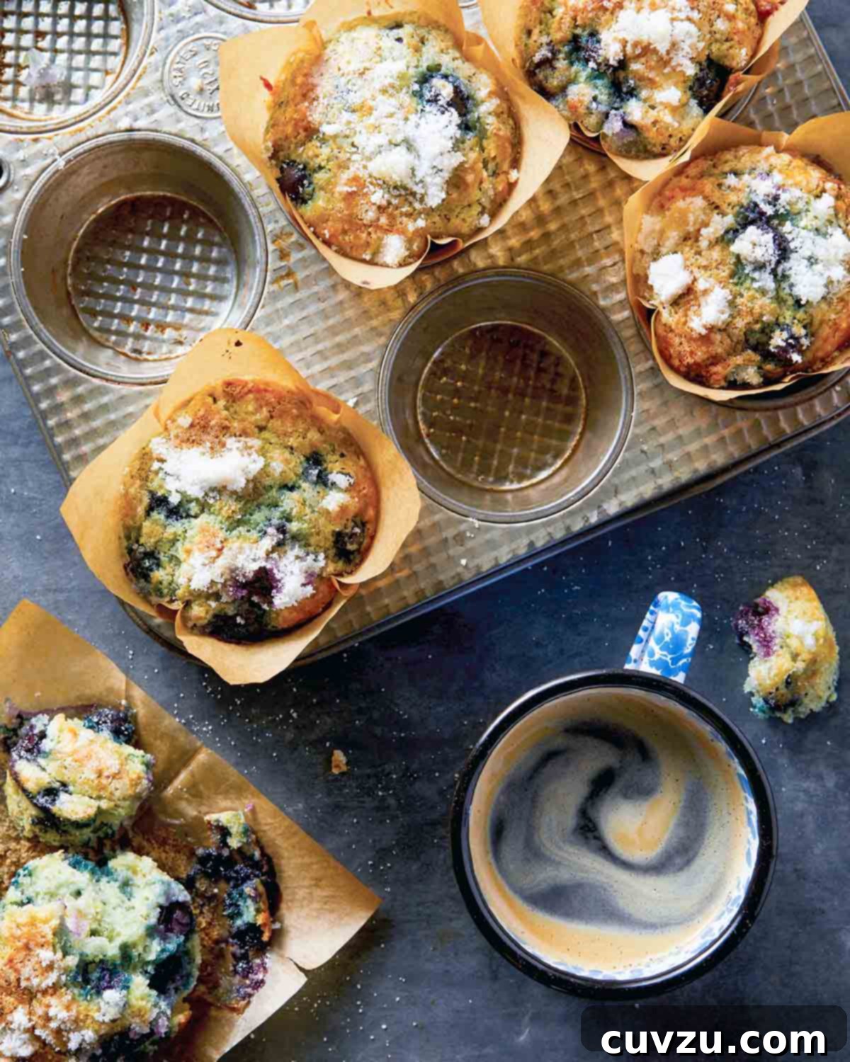 Zesty Lemon Blueberry Scones 4 A muffin tray with several blueberry muffins with sugar topping in tulip-style liners, with a halved muffin and a cup of coffee on the side.