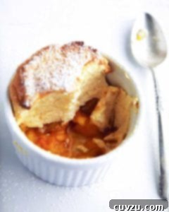 A white dish containing a sugar-dusted individual peach soufflé, with visible peach slices at the bottom.