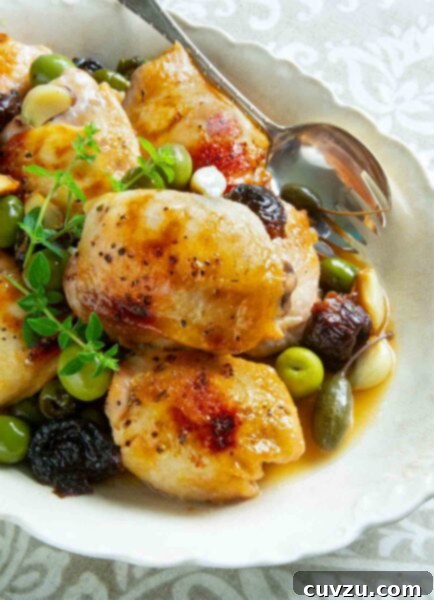 Pieces of chicken Marbella in a white serving dish with prunes, olives, and capers, garnished with fresh thyme and a serving spoon on the side.