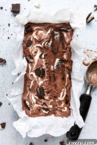 A loaf pan filled with chocolate Oreo ice cream with pieces of Oreo scattered around it and a ice cream scoop on the side.