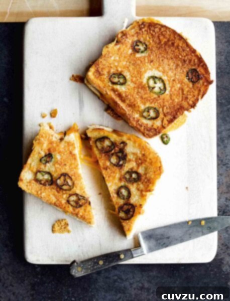 Crispy Parmesan Jalapeño Grilled Cheese 3 Two Parmesan-crusted grilled cheese with jalapeños on a white cutting board, one cut in half, with a knife lying beside them.