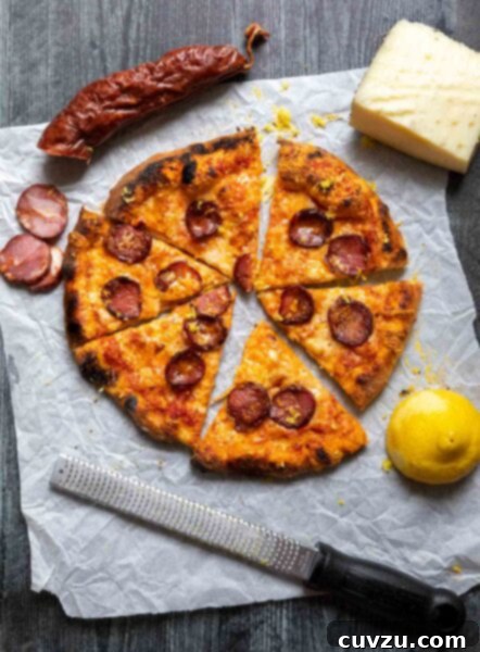 An Ooni pizza with Portuguese sausage and cheese on gray weathered wood, with chouriço, lemon, cheese, and pizza cutter.