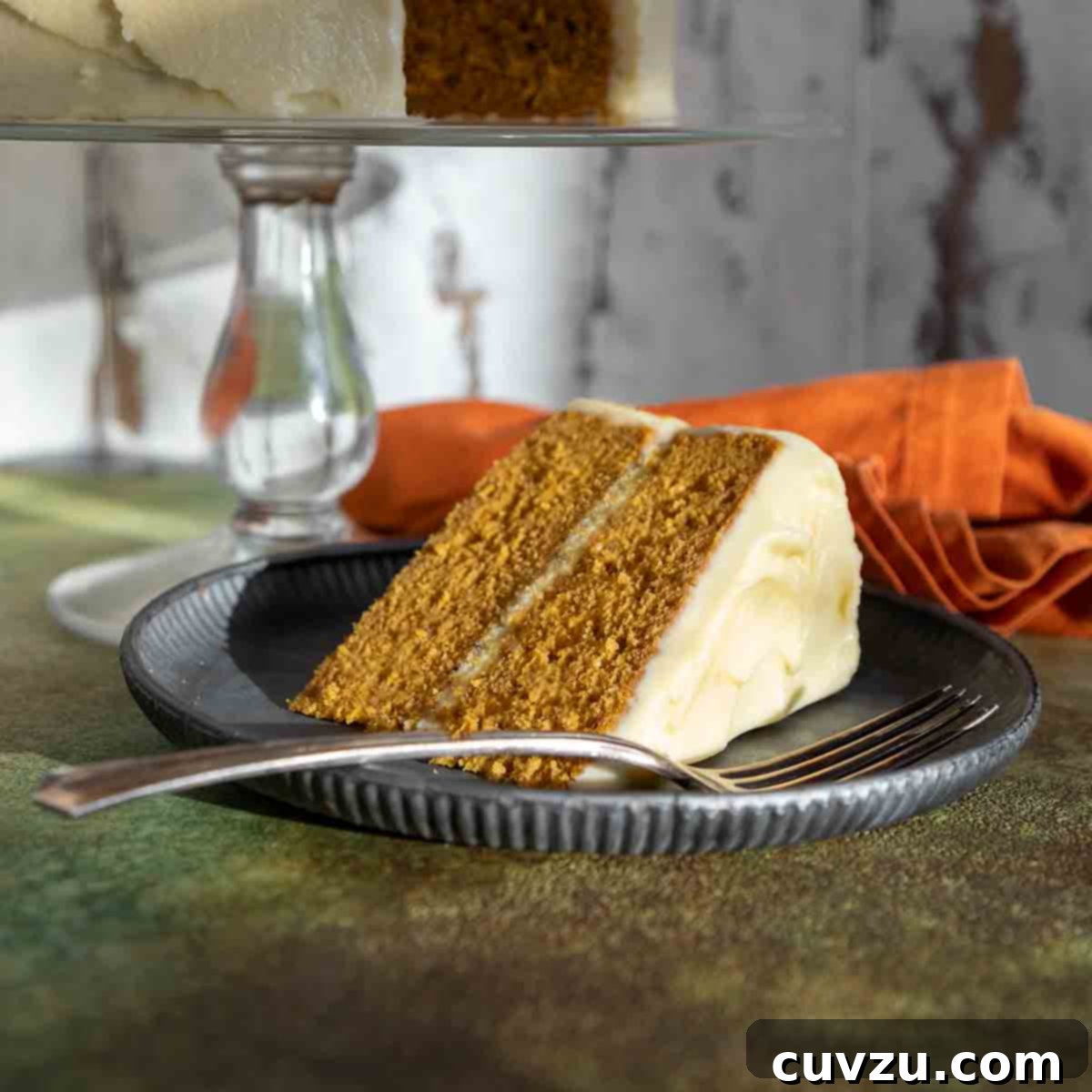 A slice of frosted pumpkin cake on a plate with the remaining cake on a stand in the background.