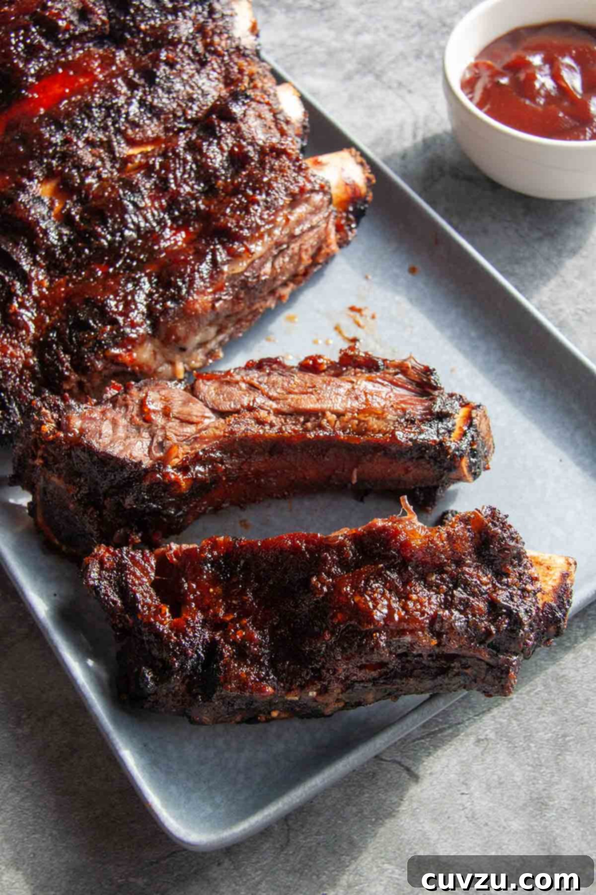 Slow-Smoked Beef Back Ribs 7 A rack of barbecued beef back ribs on a rectangular platter with a dish of barbecue sauce on the side.