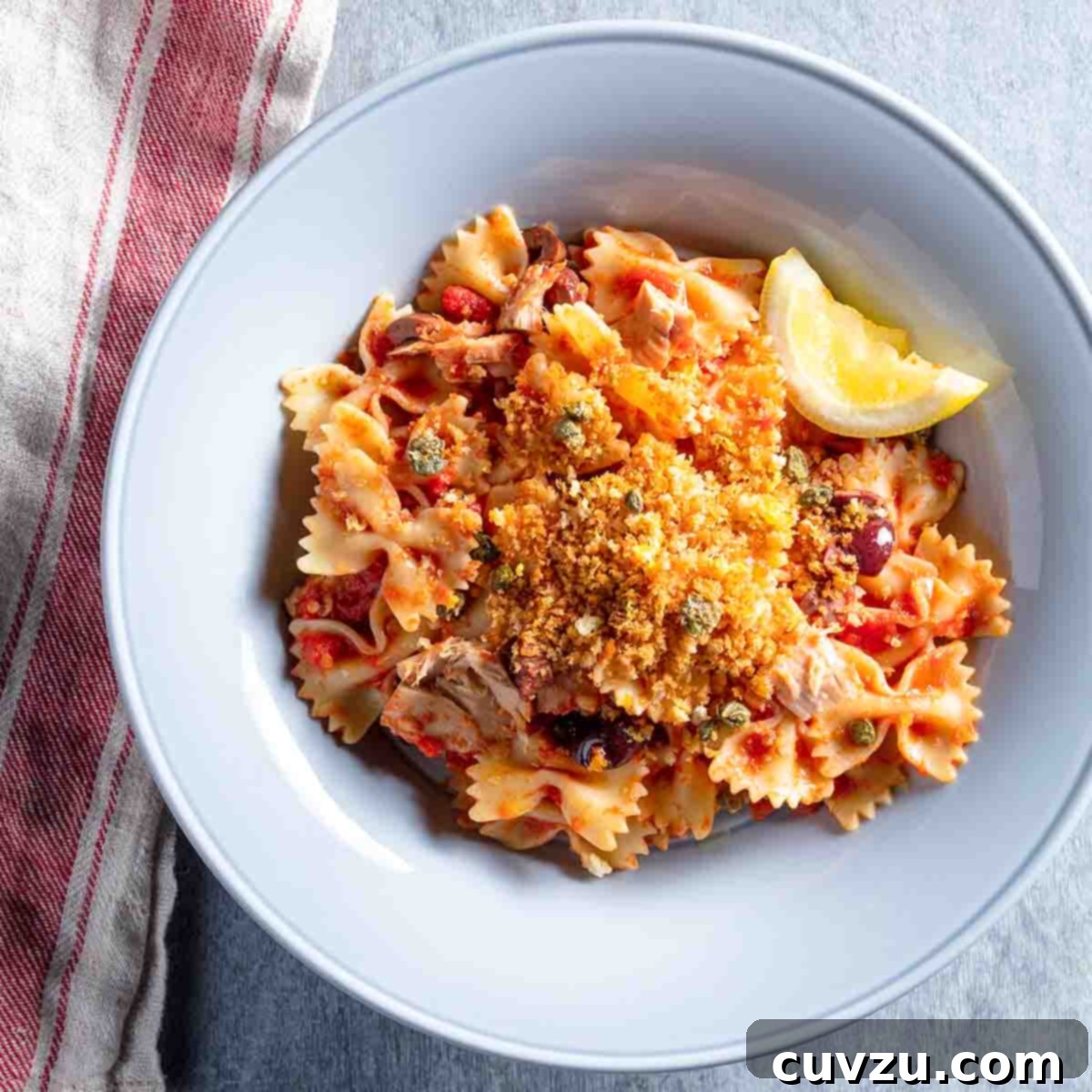 A pale blue bowl filled with farfalle, tuna, capers, tomatoes, and topped with bread crumbs and a slice of lemon, sitting on a red and white dish cloth.