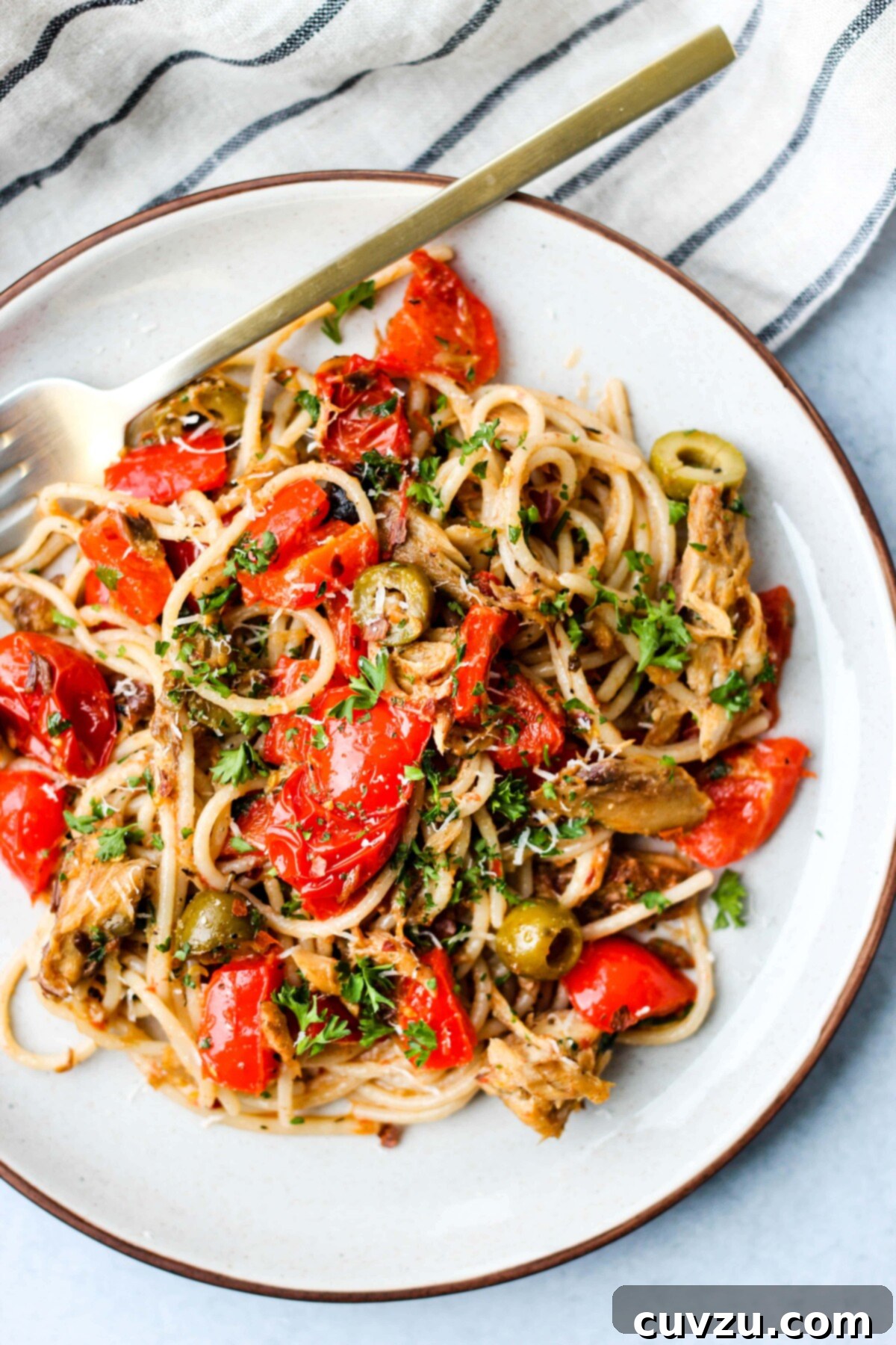 A white plate filled with pasta, canned mackerel, tomatoes, olives, and fresh parsley, with a gold fork on the side.