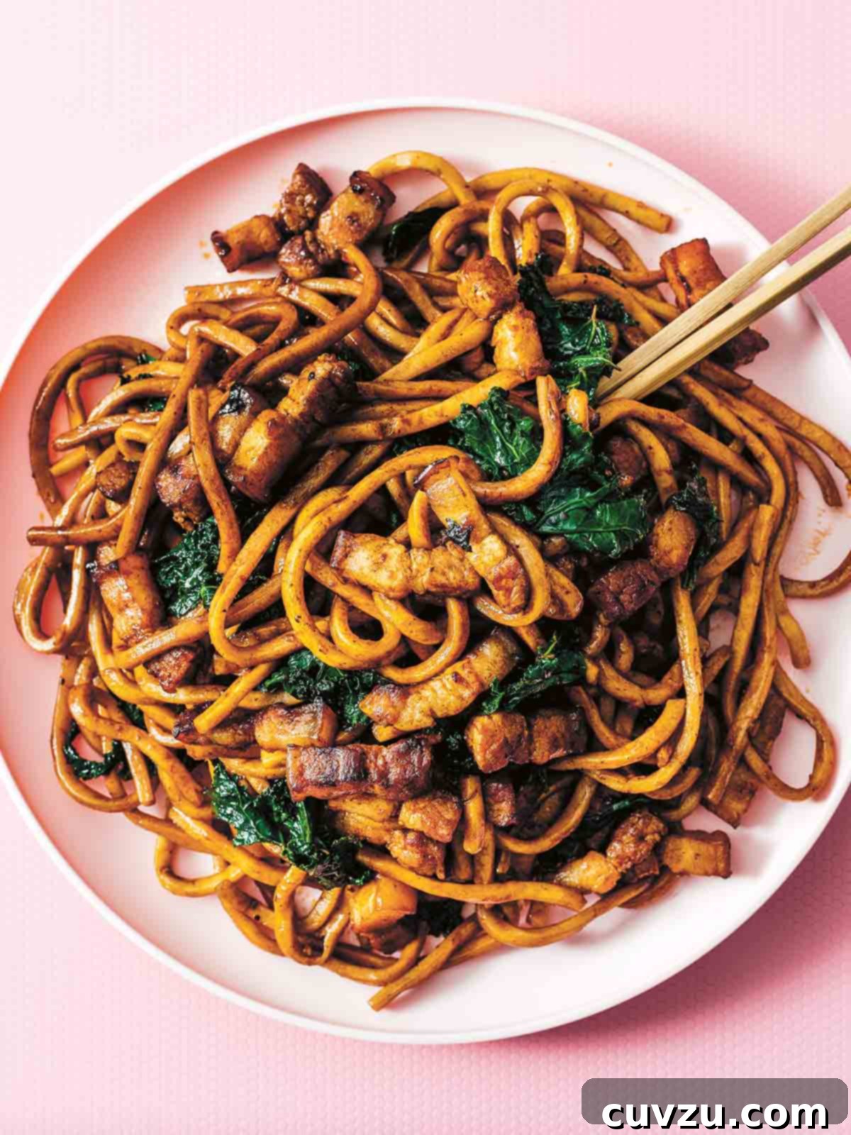 A white bowl filled with fried Shanghai noodles, crispy pork belly, and kale, with chopsticks resting on the side.