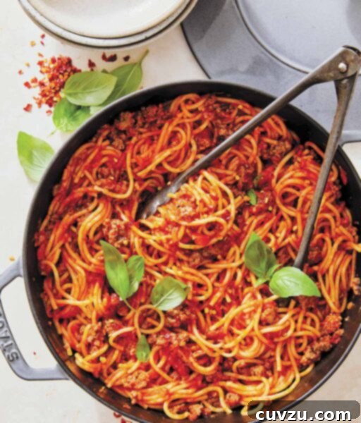 A skillet filled with spaghetti and meat sauce with a pair of tongs and two basil sprigs on top.