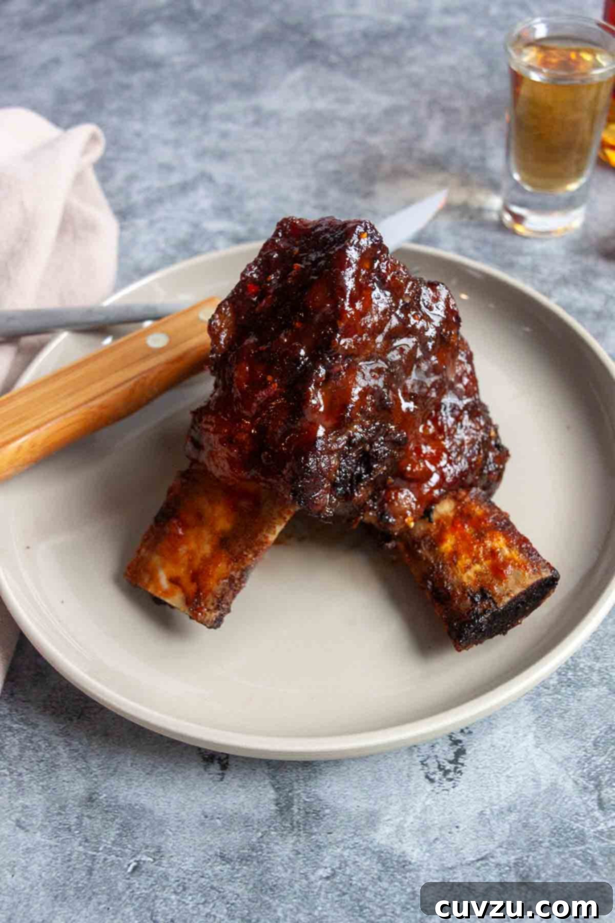 Smoky Bourbon-Glazed Short Ribs 9 Two cooked short ribs on a plate, gleaming with sticky BBQ sauce, accompanied by a shot of bourbon in the background and a fork and knife on the side, ready to be enjoyed.