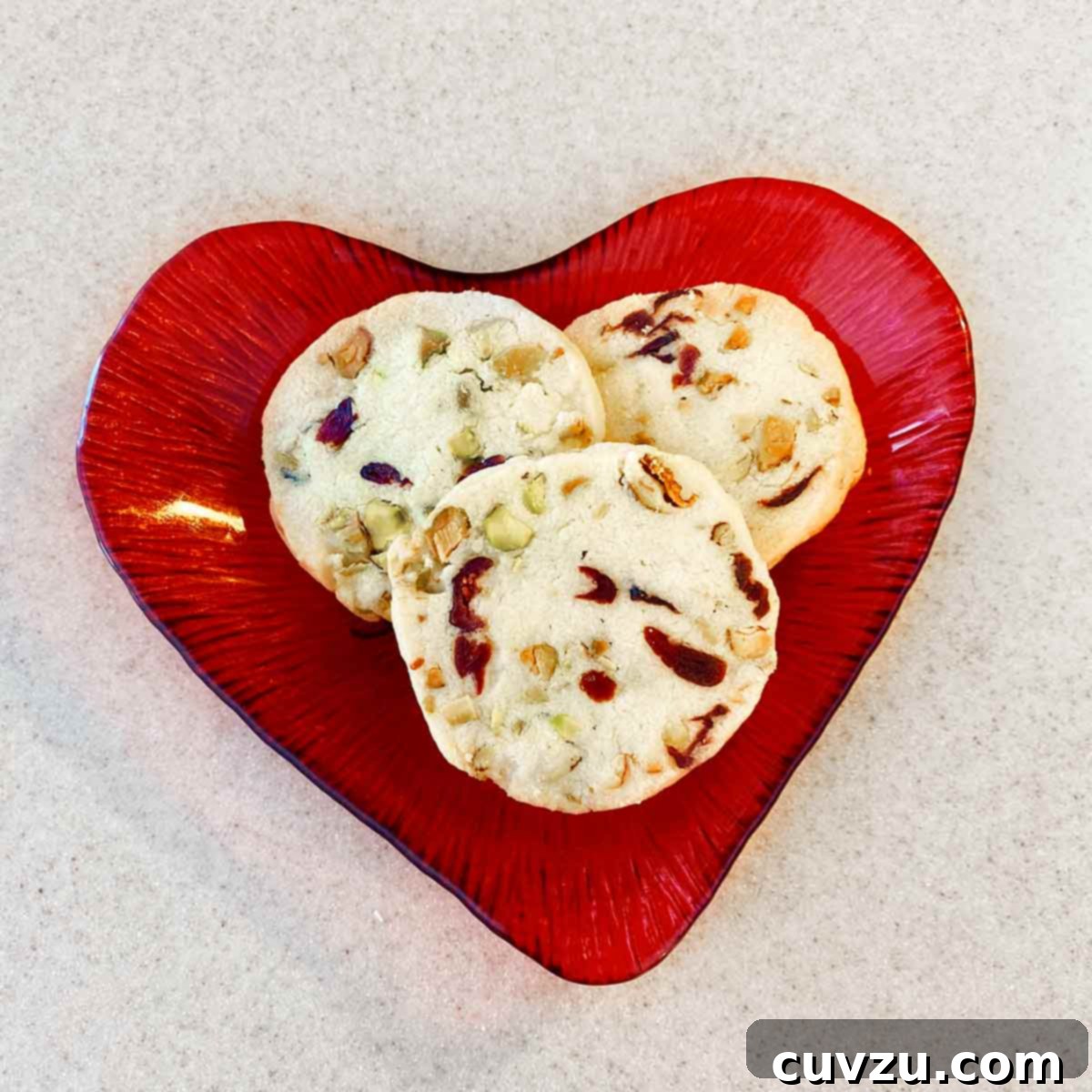 Jewel Toned Pistachio Cookies 12 Three perfectly baked cranberry pistachio cookies arranged on a heart-shaped red plate, ready to be enjoyed.