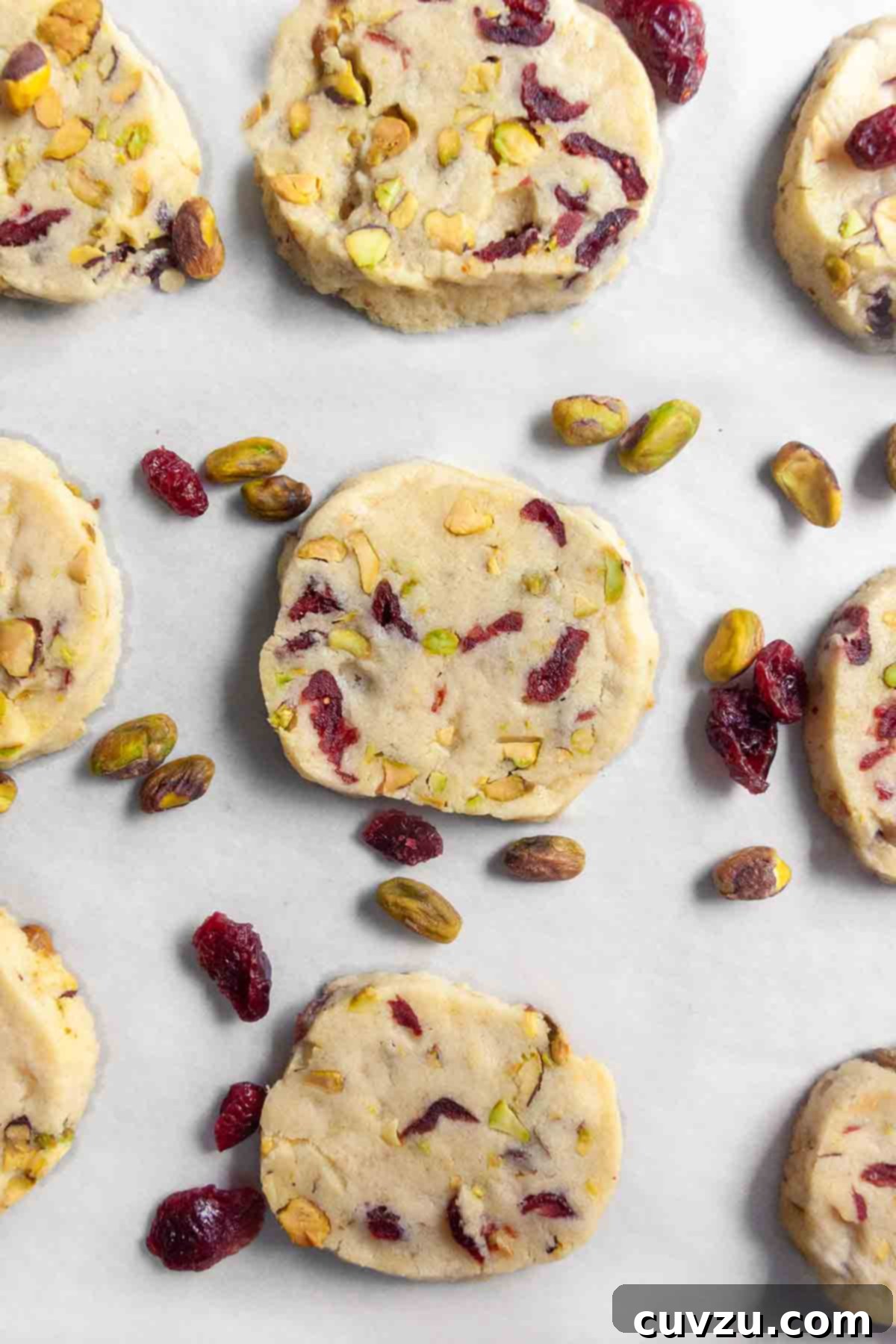 Jewel Toned Pistachio Cookies 8 A charming display of nine freshly baked cranberry pistachio cookies artfully arranged on a sheet of parchment paper, complemented by scattered pistachios and dried cranberries, highlighting their festive colors and enticing texture.