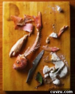 A perfectly cooked salt-baked red snapper, with its flesh carefully removed from the bones and a broken salt crust, showcasing moist, tender fish. A knife rests beside the fish skeleton, indicating the carving process.