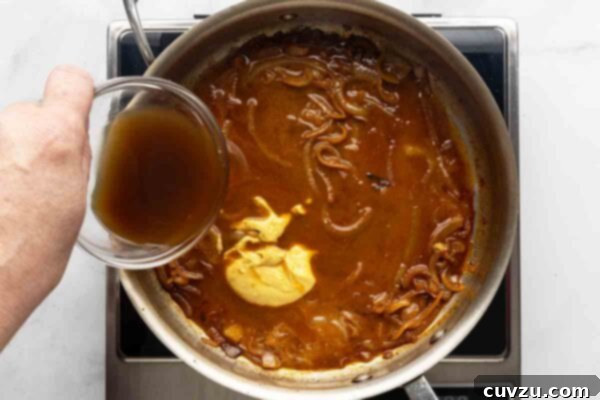 Velvety Meatball Stroganoff 16 A person adding beef broth and Dijon mustard to a onion gravy in a skillet.