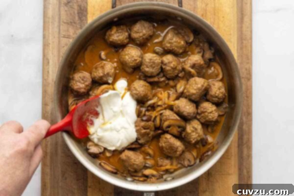 Velvety Meatball Stroganoff 18 A skillet filled with meatball Stroganoff and a spatula stirring in sour cream.