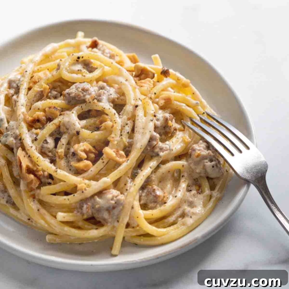 A white bowl filled with blue cheese, sausage, and walnut pasta with a fork resting on the side of the bowl.