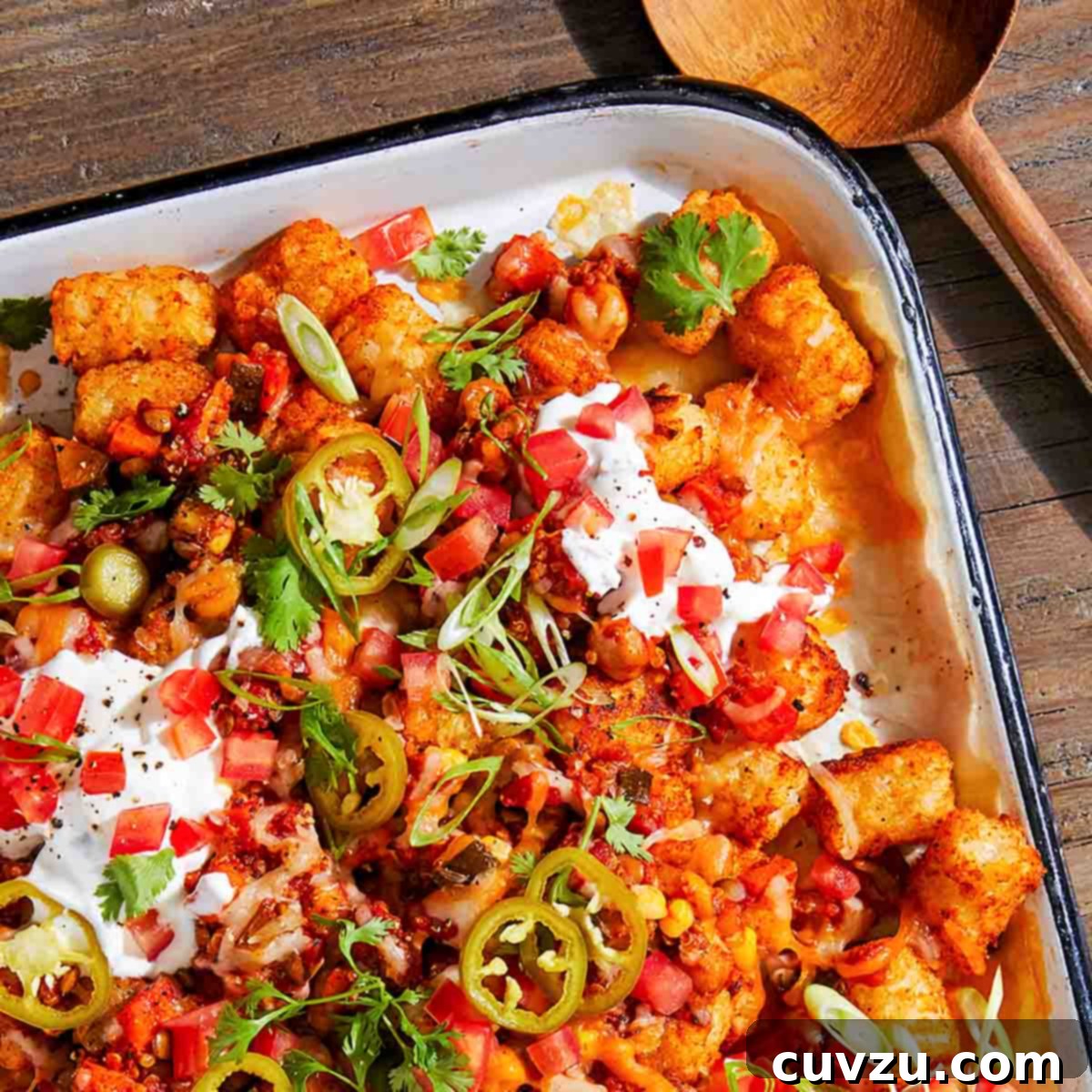A large rectangle casserole pan, filled with tater tot nachos topped with sour cream, tomatoes, sour cream, and jalapeños. A wooden serving spoon lays beside the dish.