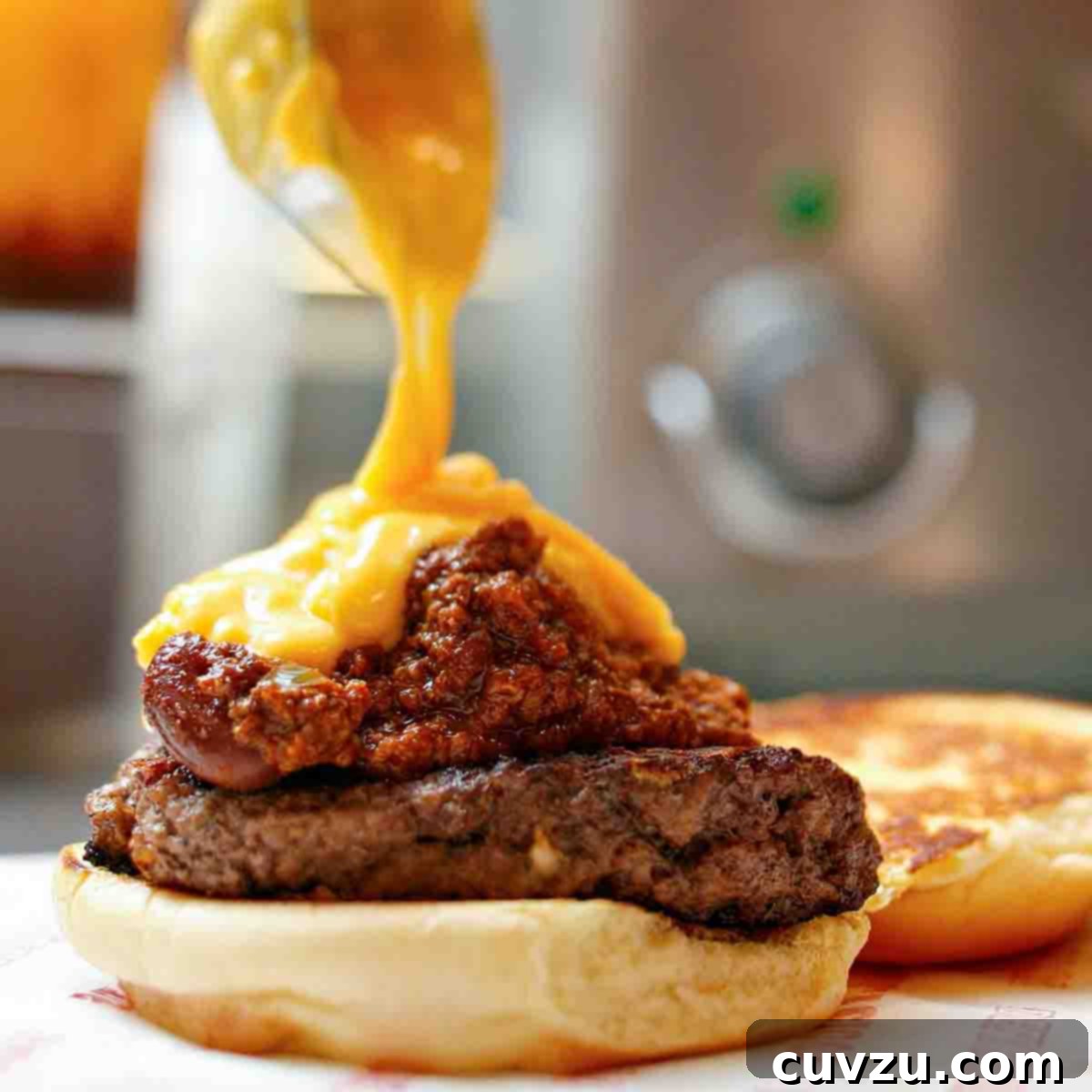 A chili cheddar burger on a bun half with cheese sauce being poured over it.