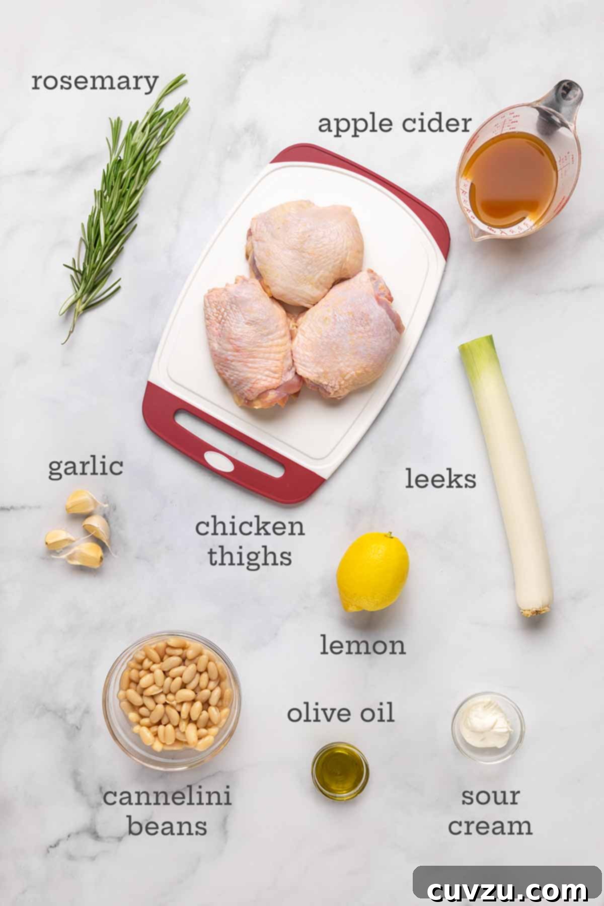 Velvety Cider-Braised Rosemary Chicken with Cannellini Beans 4 A flat lay photograph displaying all the fresh ingredients needed for rosemary chicken: bone-in chicken thighs, fresh rosemary sprigs, a bottle of apple cider, garlic cloves, sliced leeks, a bright yellow lemon, a can of cannellini beans, olive oil, and a dollop of sour cream.
