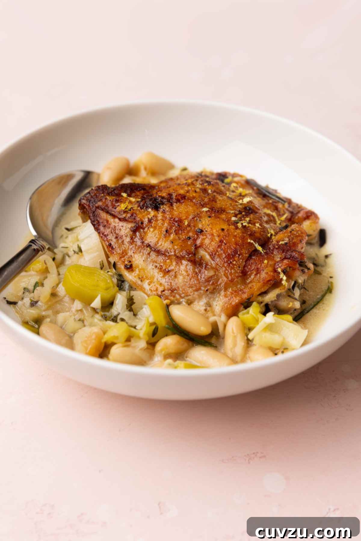 Velvety Cider-Braised Rosemary Chicken with Cannellini Beans 9 A rustic white bowl containing one perfectly crispy rosemary chicken thigh, nestled atop a generous bed of creamy leeks and cannellini beans, all coated in the flavorful cider broth.