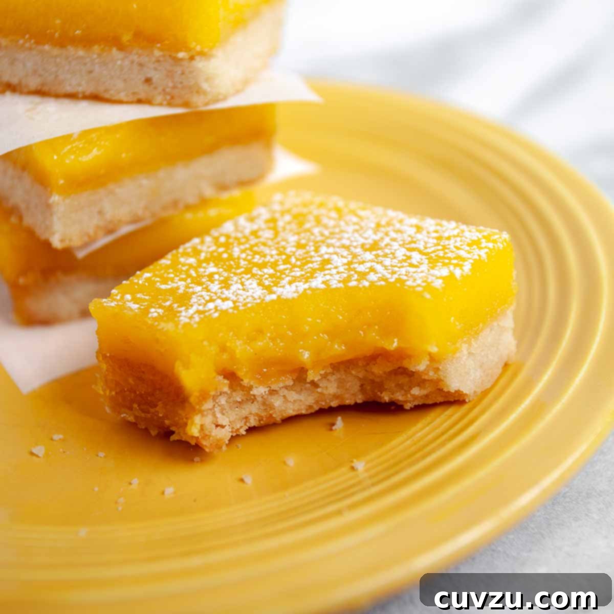 Yellow plate with four lemon bars sprinkled with powdered sugar, one with a bite take out of it.