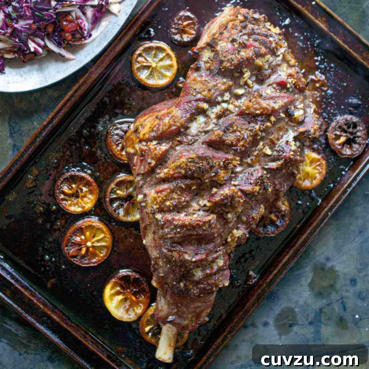 A whole roast leg of lam on a rimmed baking sheet with charred lemon slices scattered around it.