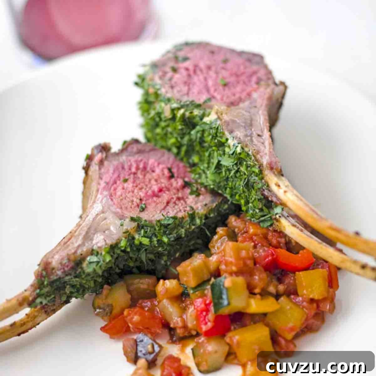 Two chops from a roasted rack of lamb with parsley, Dijon, and chives on a white plate with a cooked zucchini and tomato mixture.
