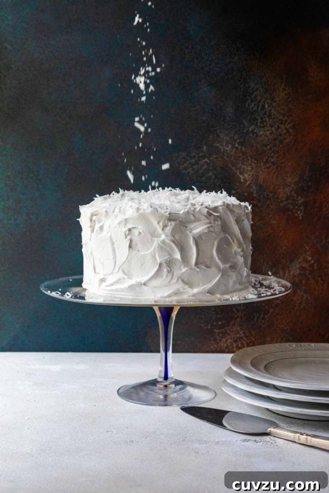 A white old-fashioned coconut cake with 7-minute frosting topped sprinkled with shredded coconut on a glass cake stand.