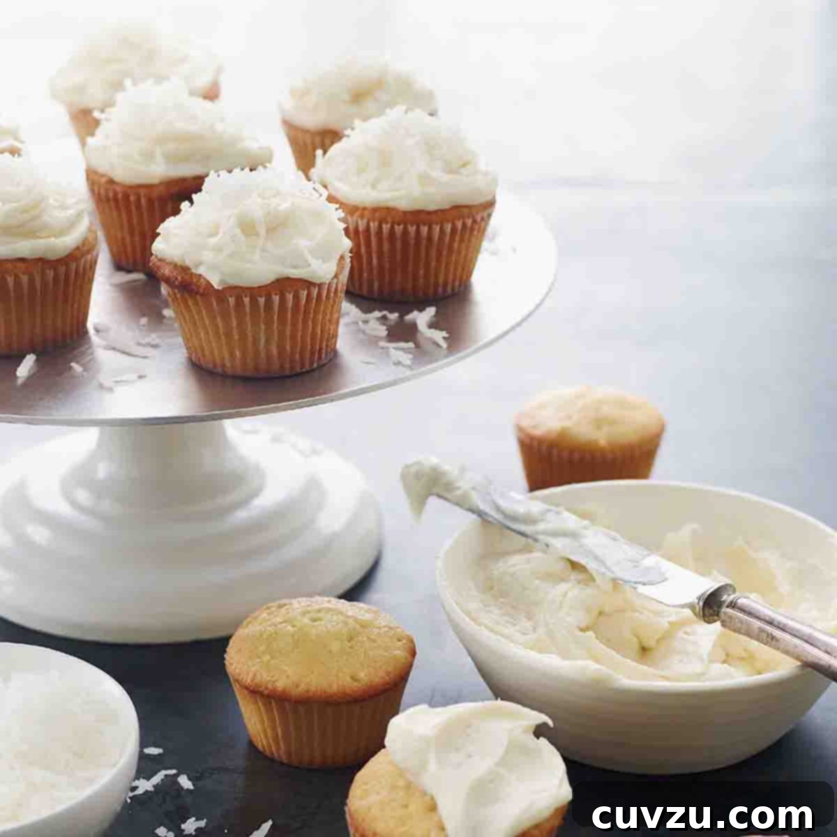 Cake stand with 7 frosted coconut cupcakes, four unfrosted cupcakes on the table, and a bowl of frosting.