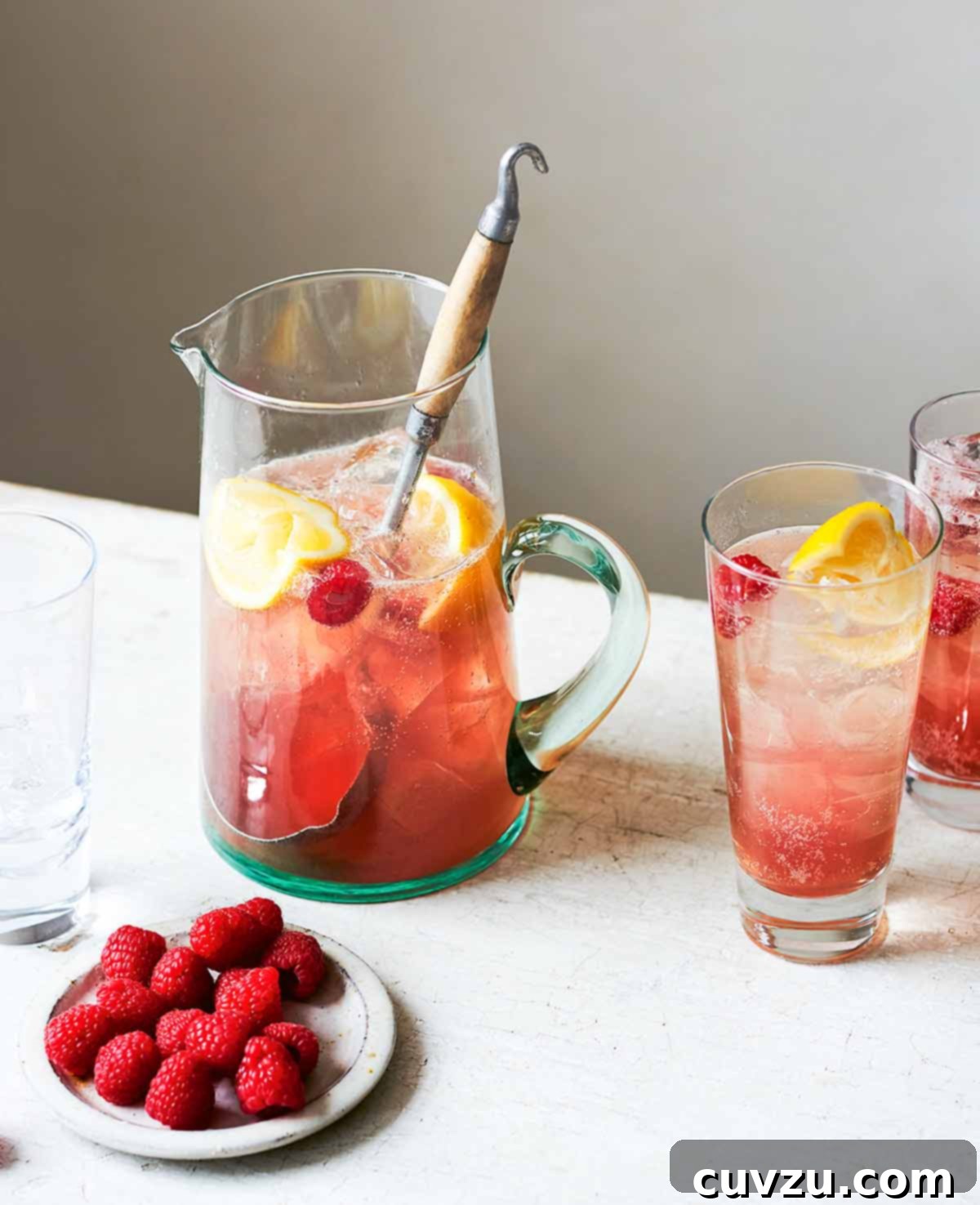 Raspberry vodka lime soda in a pitcher with a large spoon, beside two full glasses, one empty glass, and and a dish of raspberries.