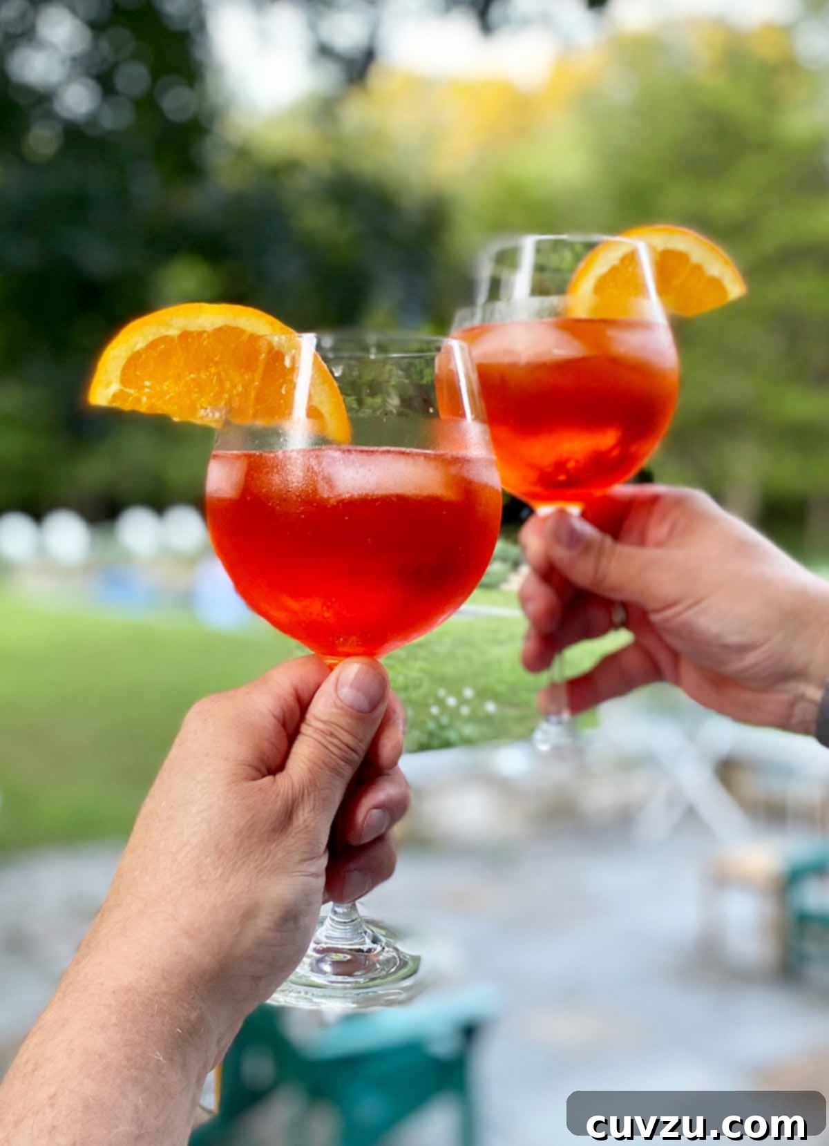 2 hands, each holding a wine glass 3 quarters full of a bubbly orange cocktail with ice and an orange slice.