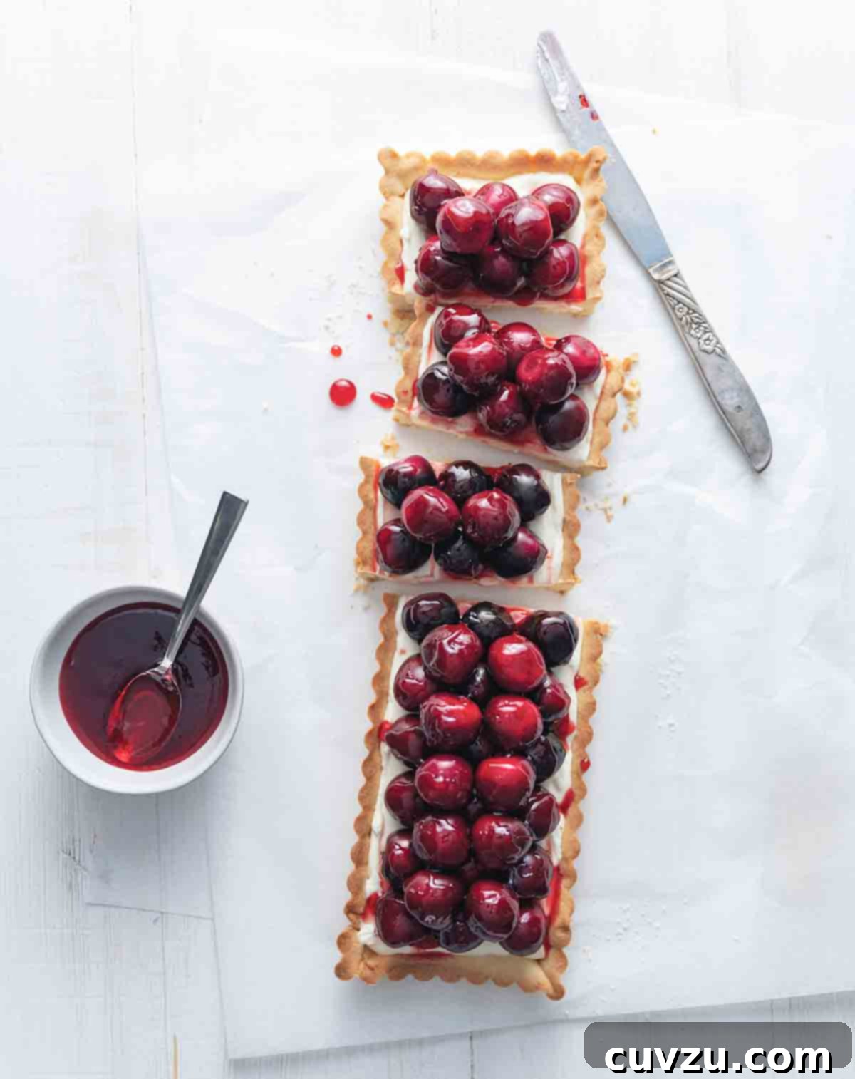 A rectangular fresh cherry tart with a creamy white filling topped with bright red cherries, displayed on a pristine white surface. A serving knife and a small bowl of jam are placed neatly beside it.