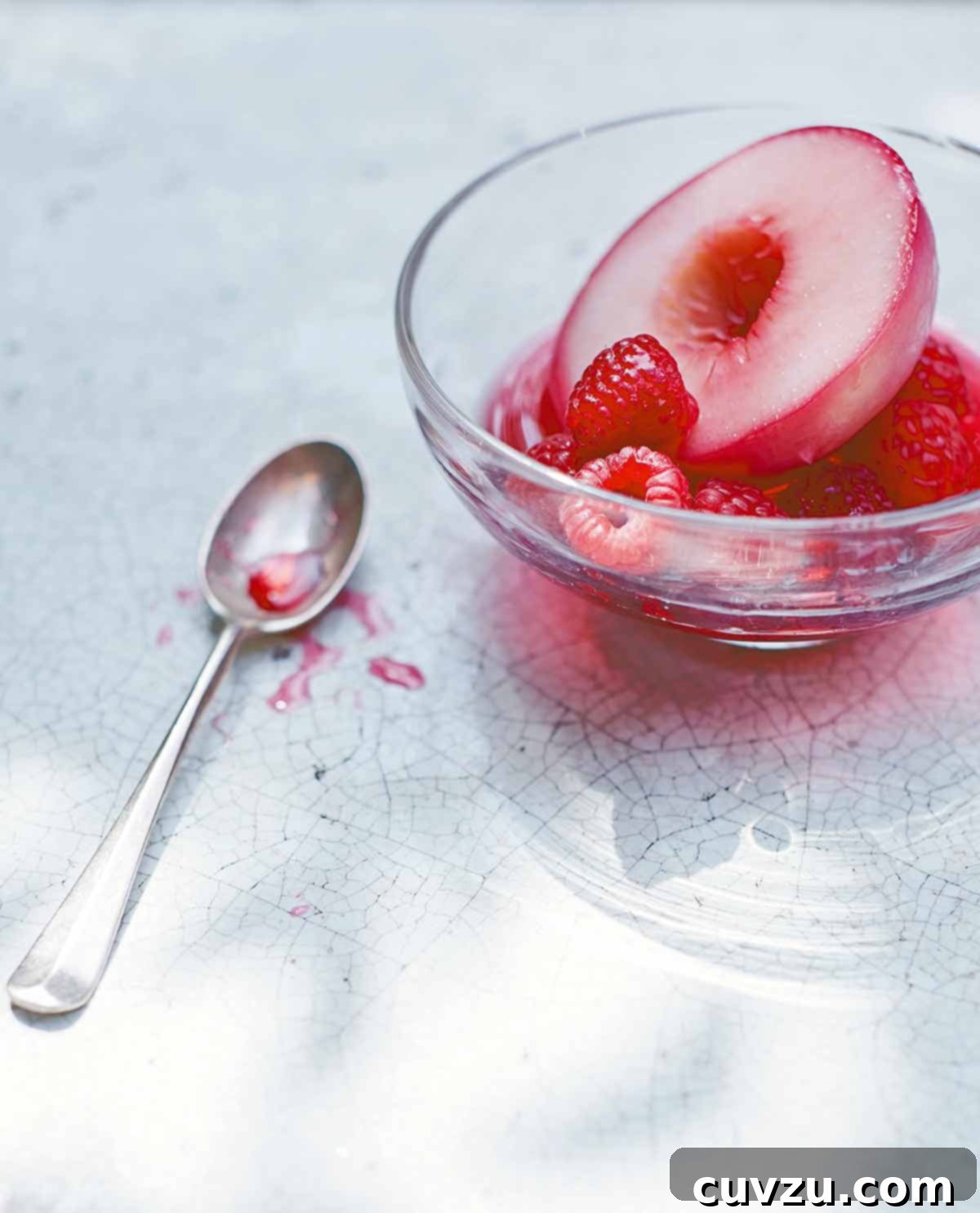 A pristine glass bowl containing a perfectly poached nectarine half, adorned with fresh, vibrant raspberries. A spoon rests casually on the side of the bowl, inviting a taste of this elegant and refreshing dessert.