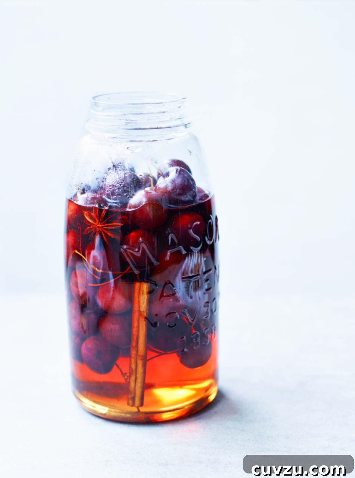 A charming mason jar largely filled with glistening pickled cherries, immersed in a spiced pickling liquid. A star anise pod and a cinnamon stick are visible within the jar, adding to its aromatic appeal.