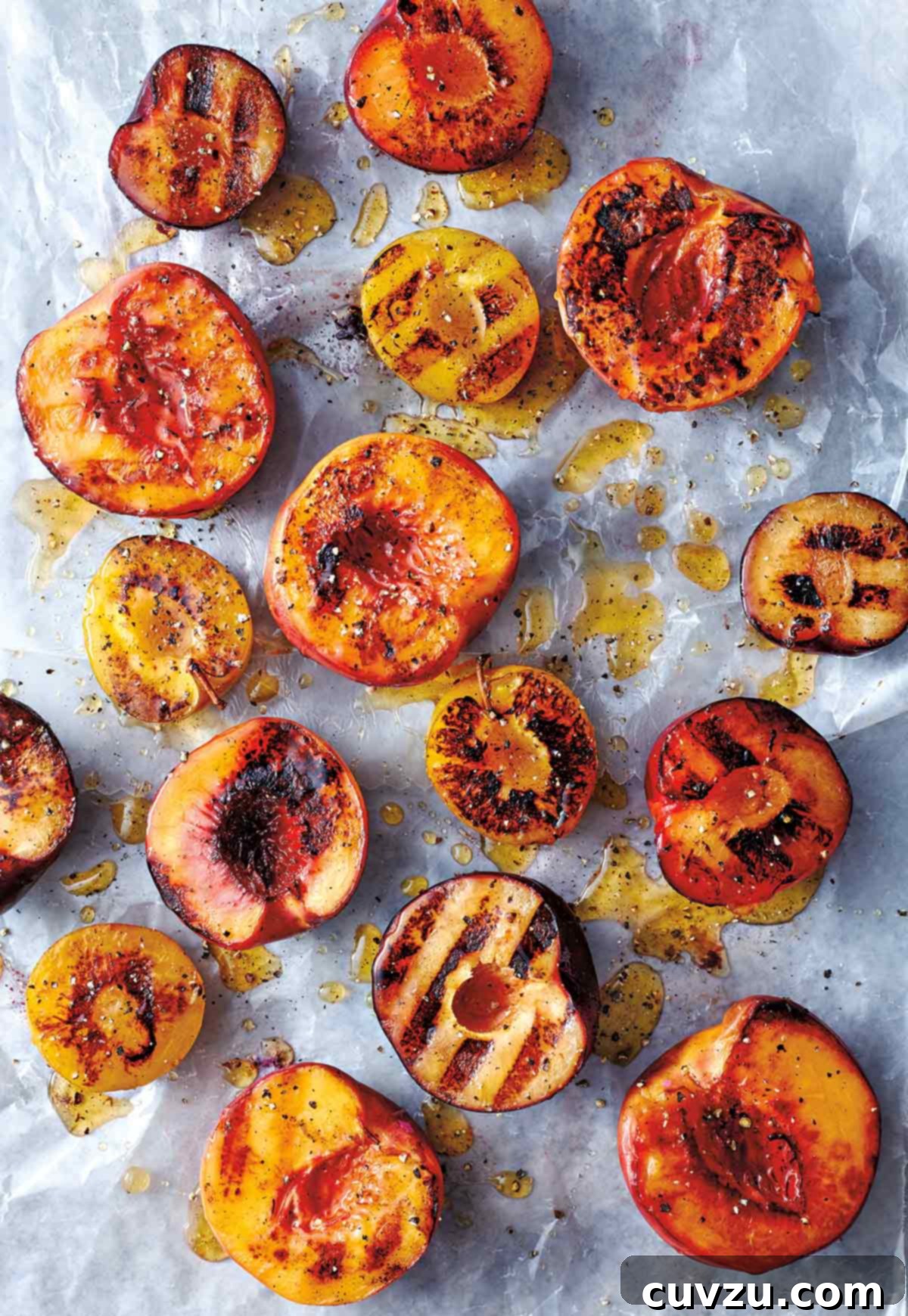 Halved grilled peaches, glistening with honey and sprinkled with black pepper, elegantly scattered on a piece of parchment paper. Their caramelized edges promise a delightful summer treat.