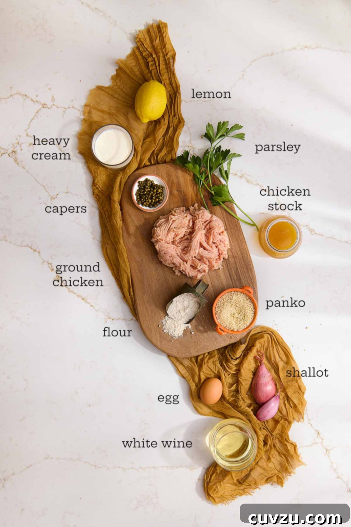 Lemon Caper Chicken Meatballs 5 Overhead view of labeled chicken piccata meatball ingredients, including ground chicken, panko, lemon, capers, heavy cream, white wine, chicken stock, flour, egg, shallot, and parsley arranged on a wood board with a golden cloth beneath.