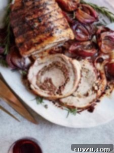 A tied pork loin in the style of Porchetta on a white platter with cooked red onions and rosemary.