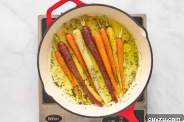 Orange juice and water poured over carrots and rosemary in a skillet, ready for seasoning.