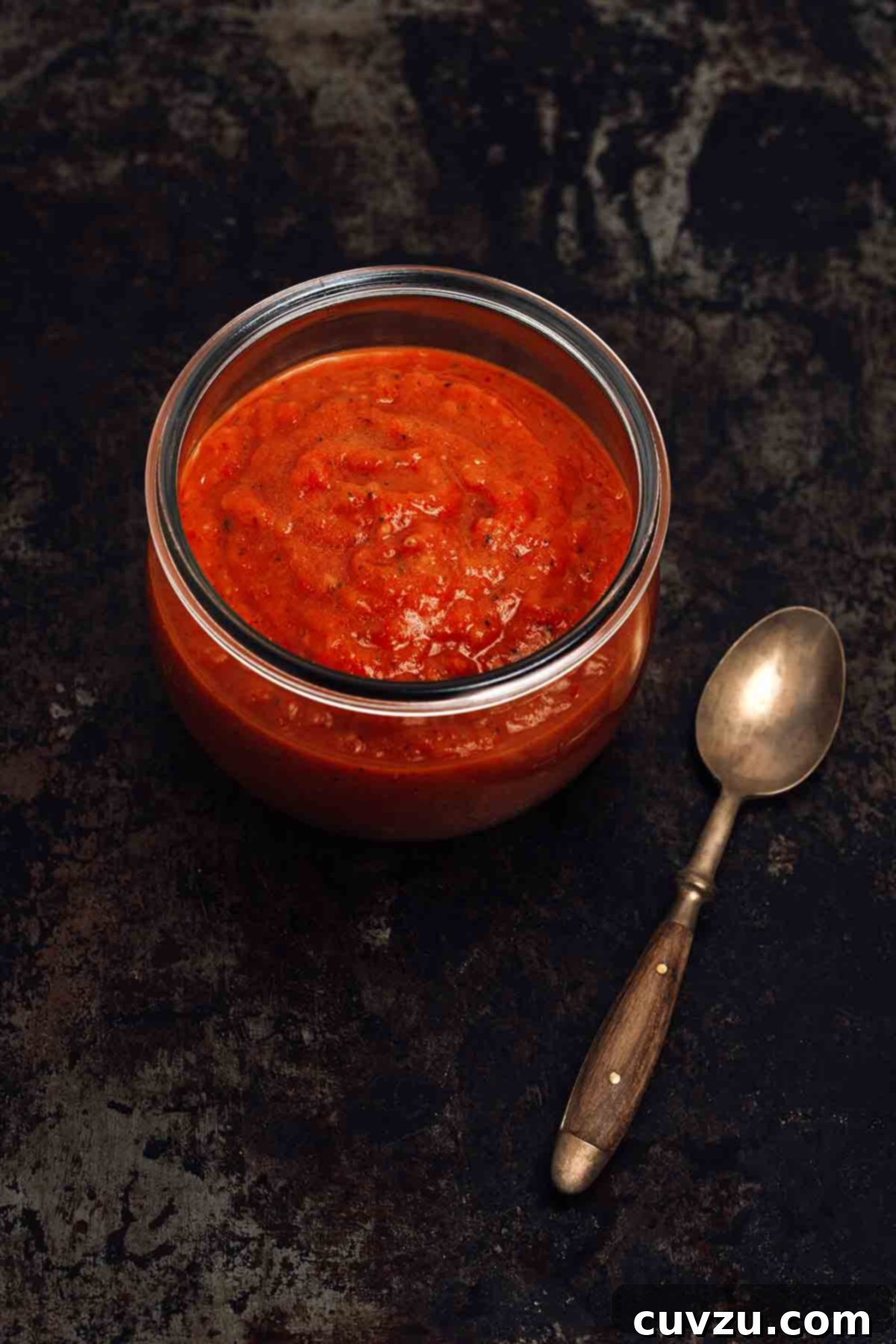 Small-Batch Sriracha Fire 3 A beautifully textured jar of rich red homemade sriracha hot sauce on a sleek black background, with a spoon resting nearby.