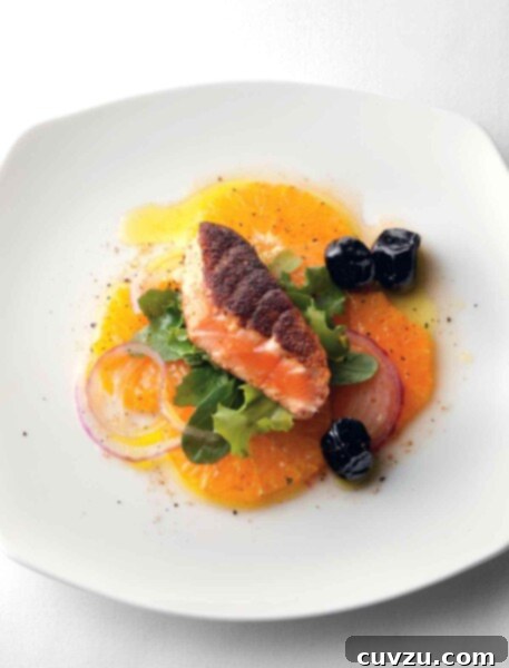 A white plate with a piece of pan-seared Moroccan salmon on a bed of orange slices, red onion, and greens with three black olives on the side.