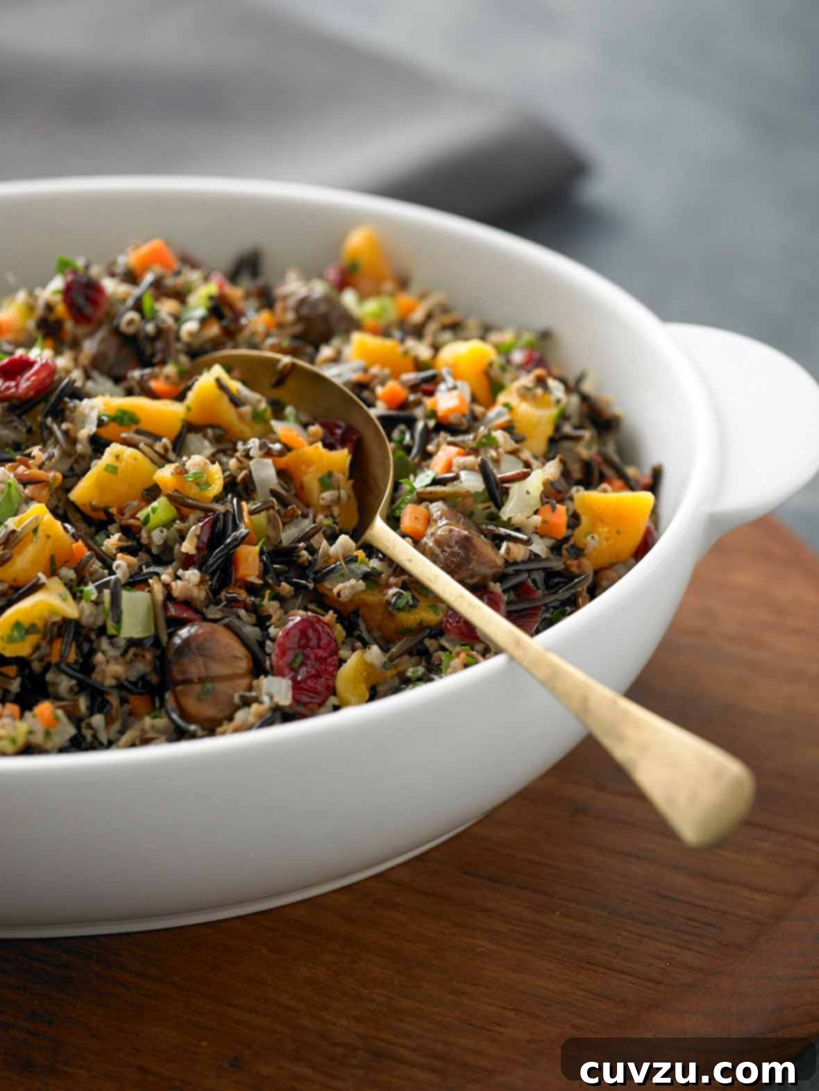 A beautifully presented white bowl of wild rice, roasted chestnuts, and cranberries, garnished with fresh parsley, with a gold spoon resting inside.