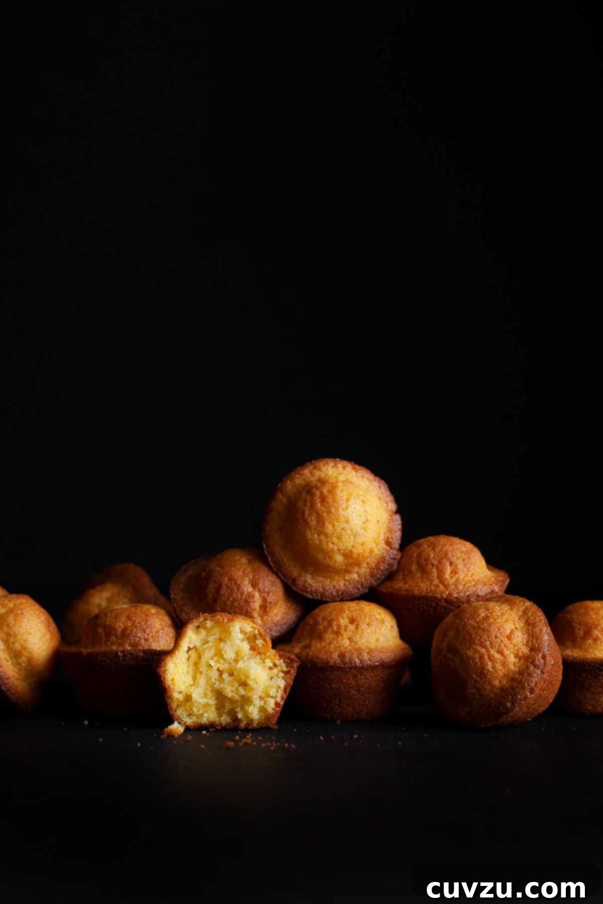 Several Portuguese mini lemon-orange cakes, some whole and one torn in half to reveal its tender crumb, scattered on a dark background.