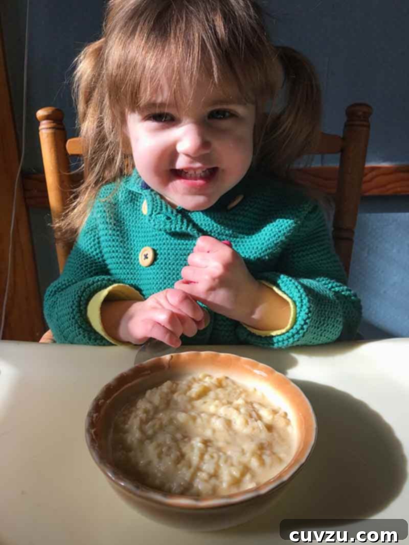 Silky Comfort Pastina 2 A young girl seated at a table, holding a spoon with a bowl of pastina in front of her, embodying pure childhood joy.